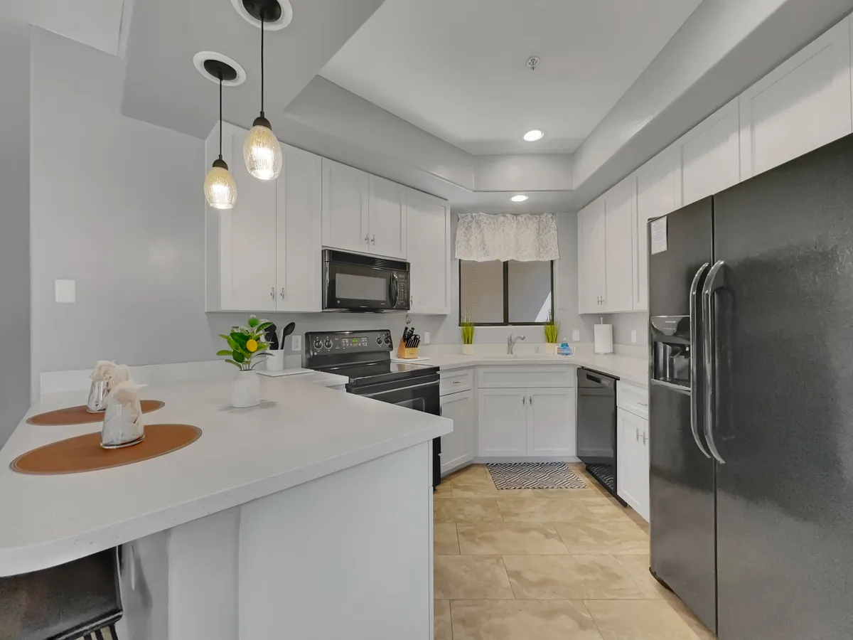 A modern kitchen featuring white cabinetry, black appliances, pendant lighting, and a decorative window treatment. A small potted plant adds a touch of greenery, and the layout is open, airy, and inviting. The visual focus is on the kitchen’s functionality and stylish design, with plenty of counter space for meal prep.
