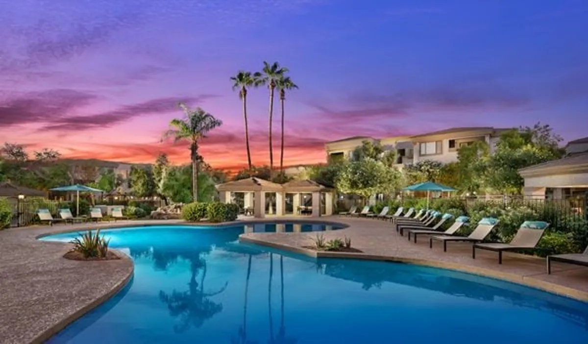 A vibrant exterior view of a sparkling swimming pool surrounded by palm trees and lounge chairs, set against a striking sunset sky. The inviting scene radiates a resort ambiance, ideal for sunny afternoons or evening relaxation.