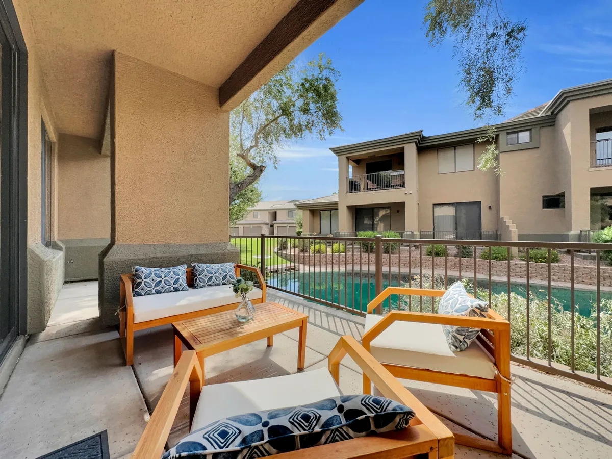 This image showcases a serene exterior seating area adorned with decorative cushions and a small vase with flowers, all overlooking a sparkling swimming pool. Manicured landscaping provides a lush backdrop, creating a tranquil environment for relaxation and socializing.