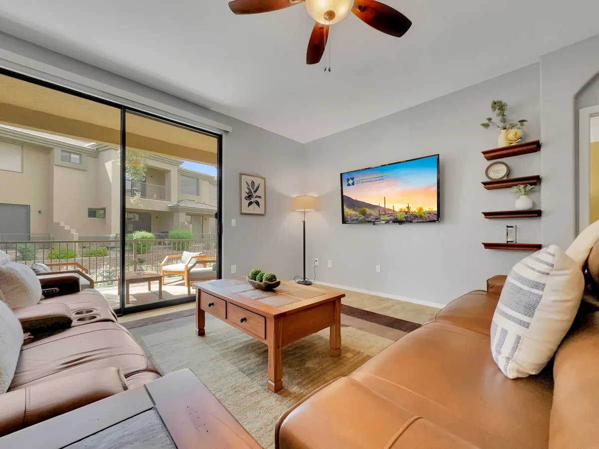 A bright, open living room featuring a comfortable leather sofa, a wooden coffee table, lush indoor plants, a television with a scenic view, and large windows allowing natural light to flood in. The space is arranged for both relaxation and gathering, highlighting the home's welcoming atmosphere.