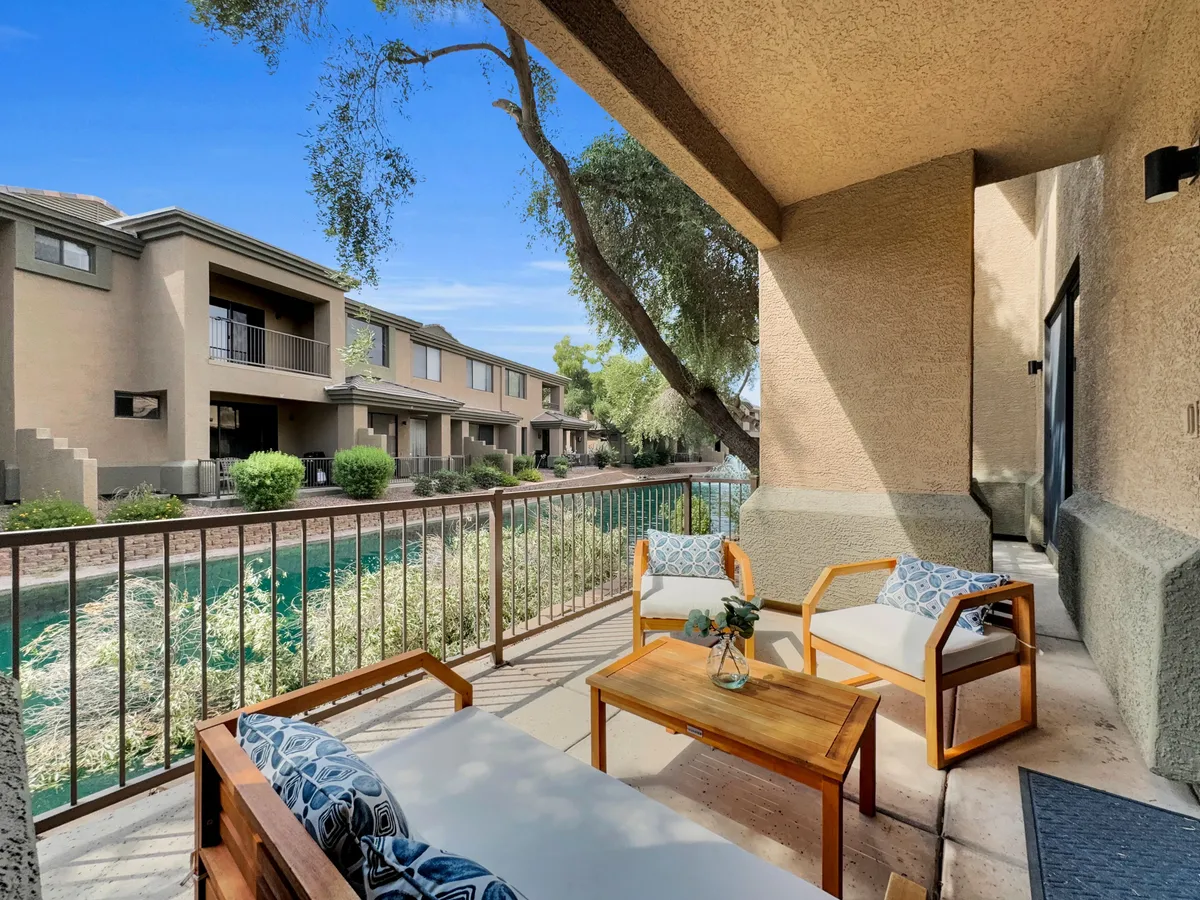 The image showcases an inviting outdoor seating area with decorative throw pillows arranged on comfortable chairs, a small table with a vase, and vibrant greenery in the background. The scene is bathed in natural light, creating a serene atmosphere perfect for relaxing or socializing while enjoying views of the surrounding landscape.