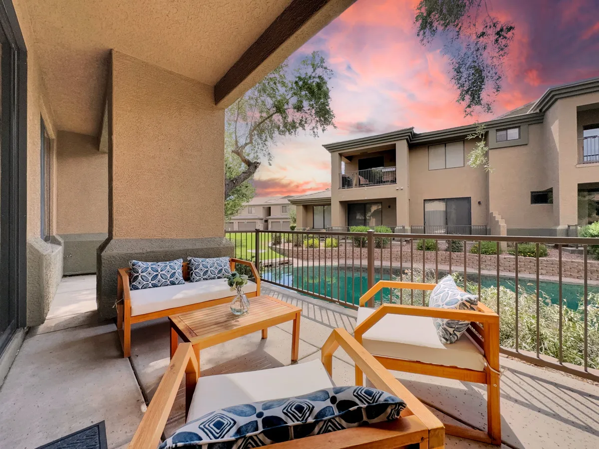 A stylish outdoor lounge area featuring comfortable seating with decorative cushions and a flower vase, set against the backdrop of a shimmering pool at sunset. The scene is tranquil, suggesting moments of relaxation, conversations, or solo reflection as the sun sets.