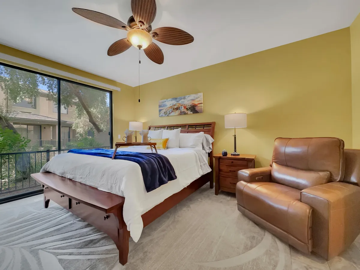 A comfortable bedroom featuring a large bed, a window with a view, a recliner by the side, and a nightstand. The room is bathed in natural light, offering both a sense of calm and a versatile layout for guests or family.