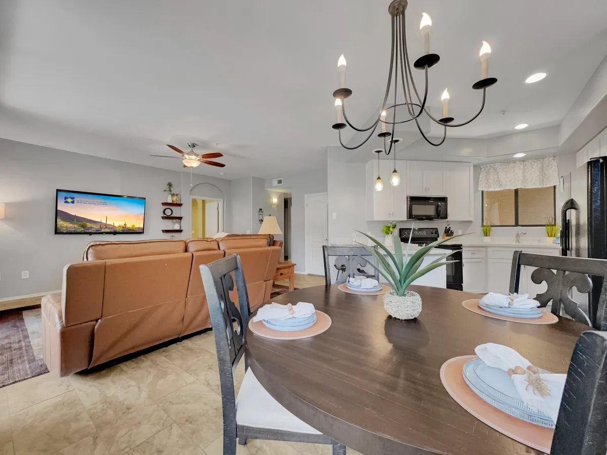 A cozy dining area with a dining table set for a meal, a chandelier overhead, and a sofa arrangement in the background. The window provides scenic views, and the table setting suggests a space ideal for family meals or entertaining guests.
